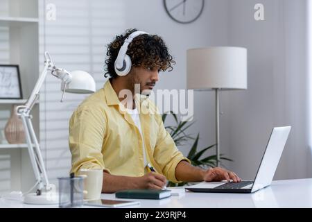 Young man concentrating on his work at home, using a laptop and headphones. Suitable for remote work, freelancing, or e-learning concepts. Stock Photo
