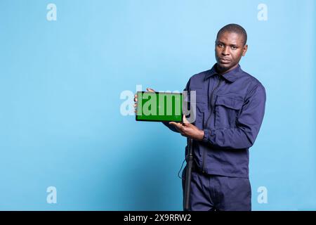Security guard using a tablet to present an isolated greenscreen on camera, wearing his work uniform and smiling. Confident agent showing mockup screen on device in studio. Stock Photo