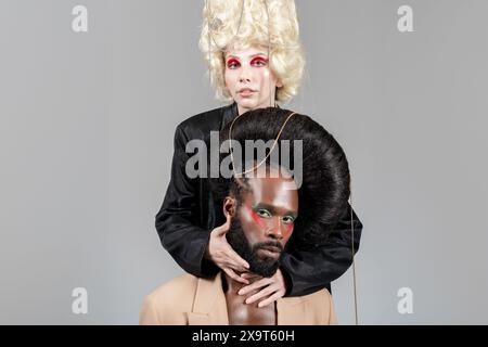 Confident gay man and woman, in vintage royal wigs and bold makeup, posing for studio portrait, looking directly at camera. Two LGBT friends in studio shot over light background. Stock Photo