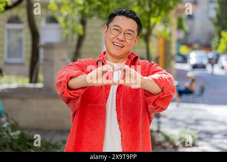 I love you. Asian middle-aged man makes symbol of love, showing heart sign to camera express romantic feelings, express sincere positive feelings outdoors. Charity, gratitude, donation. on city street Stock Photo