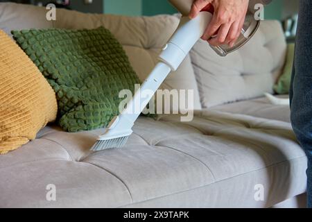 Woman is using portable cordless vacuum cleaner to clean surface of sofa in living room. House cleaning with handheld hoover. Household chores Stock Photo