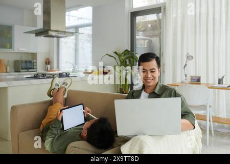 Smiling man working on laptop from home when son drawing on tablet screen Stock Photo