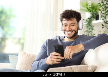 Happy man wearing headphone listening music using phone sitting on a couch at home Stock Photo