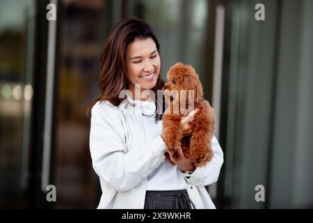 Beautiful happy dark haired woman holds and hugs small dog poodle, smiling against gray background. Moment of joy and affection between the woman and Stock Photo