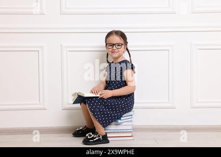 Cute little girl in glasses sitting on stack of books near white wall Stock Photo