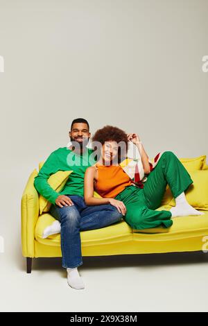 Happy African American friends in vibrant clothes sitting on a yellow couch against a grey background. Stock Photo