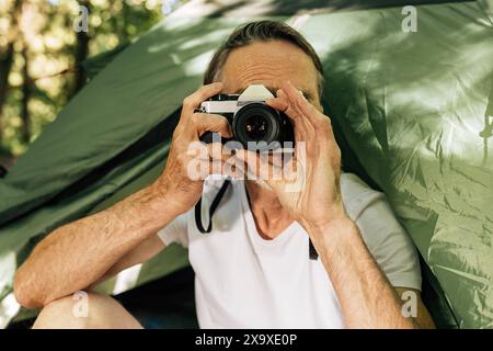 Senior male photographing while sitting in the forest during a hike. Mature man with a film camera sitting a tent outdoors. Senior male photographing Stock Photo