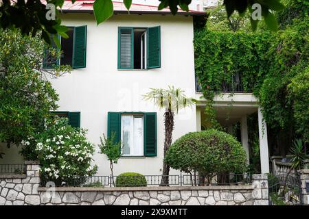 A charming two-story house with green shutters surrounded by lush greenery and well-manicured bushes Stock Photo