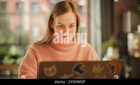 Beautiful Female Working on Laptop Computer while Sitting at a Table in Stylish Cozy Loft Living Room. Freelancer Working From Home. Browsing Internet, Using Social Networks, Being Productive. Stock Photo