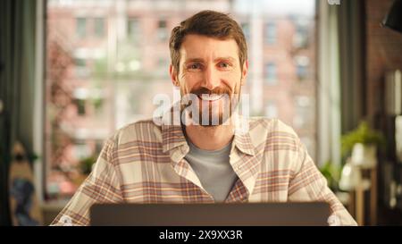 Portrait of a Happy Handsome Caucasian Man Working on Laptop Computer in Stylish Cozy Living Room. Freelancer Working From Home. Man Posing for Camera, Charmingly Smiling. Stock Photo