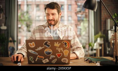 Creative Bearded Man Working on Laptop Computer while Sitting at a Table in Stylish Cozy Loft Living Room. Freelancer Working Remotely. Browsing Internet, Using Social Networks, Writing Project. Stock Photo