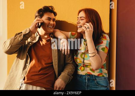 Two friends share a fun moment talking on the phone, laughing together while leaning against a colorful wall. Stock Photo