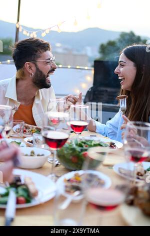 Laughing Caucasian young couple sitting at table having fun during celebration barbecue outdoors. Stock Photo