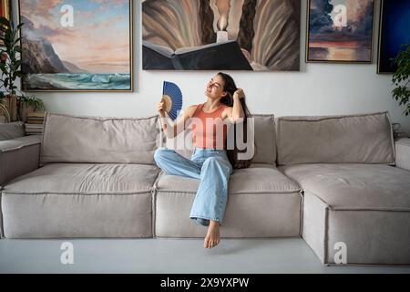 Glad woman successfully coping with abnormal heat, blowing with paper fan, sitting on couch. Stock Photo