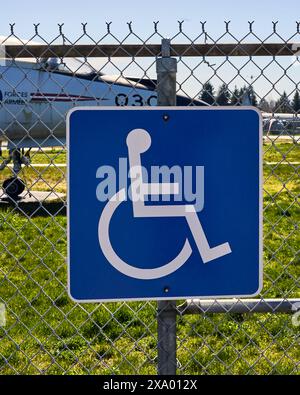 Blue and white sign with the symbol for a person in  wheelchair on a chain link fence indicating a park is handicap accessible. Stock Photo