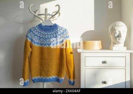Photo of a yellow blue white icelandic lopapeysa sweater with traditional Nordic pattern hanging on a rack against a white background in morning sunli Stock Photo