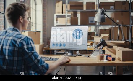 Young Man Using Desktop Computer with a Trend Analysis Charts Display. Sales Manager and Employee Working in Distribution Warehouse with Cardboard Boxes. Over the Shoulder Stock Photo