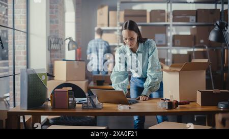 Small Business Owner Works on Laptop Computer while Standing at Her Desk in Warehouse. Female Employee Packing an Online Order in the Room with Shelves Full of Cardboard Box Parcels. Stock Photo
