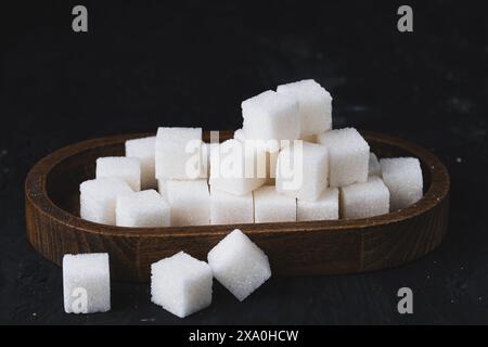 White cubes of refined sugar in a wooden plate. Sugar cubes in a wooden bowl on black background. Stock Photo