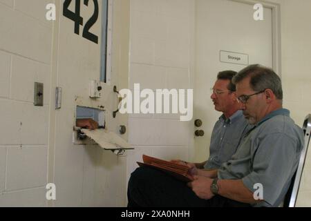 Two male individuals are seen sitting in a prison cell, reading documents, in front of a barred door. Correctional Stock Photo
