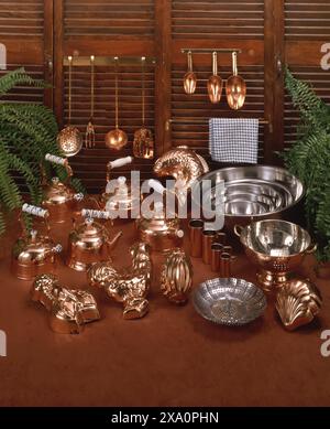 Assorted vintage copper kitchenware and utensils displayed on wooden surface with shutters backdrop, featuring kettles, molds, bowls and ladles Stock Photo