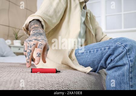 Young tattooed man sitting on bed and using disposable electronic cigarette at home Stock Photo