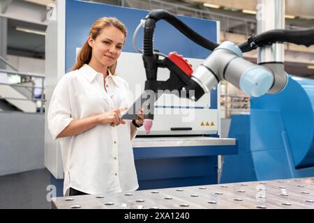 Young adult female entrepreneur controls robotic arm in startup factory using wireless technology. Stock Photo