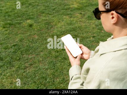 Woman stands on the green lawn and using her smartphone. Mockup blank screen of mobile phone in female hands. Stock Photo