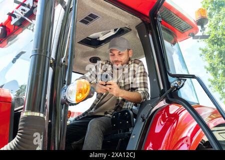 Male farmer sits on tractor, uses mobile phone and smiling. Stock Photo