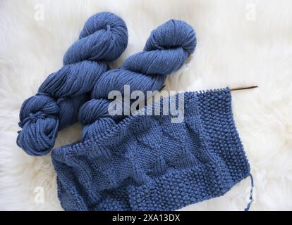 flat lay photo of a hand knitted blue merino blanket on the needles with two yarn skeins on a white lambskin fur Stock Photo