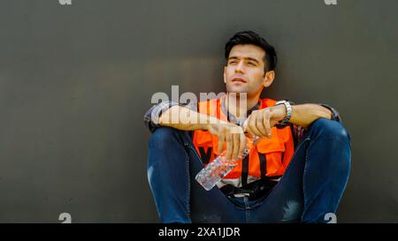 A construction worker holding a water bottle, resting against a gray wall Stock Photo