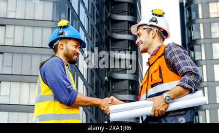 The two construction workers in hard hats shake hands outdoors with blueprints. Stock Photo