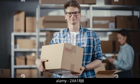 Portrait of a Successful Young Small Business Owner Smiling, Looking at Camera. Caucasian Male Holding a Cardboard Box in a Warehouse Storeroom with Orders Ready for Shipment. Stock Photo