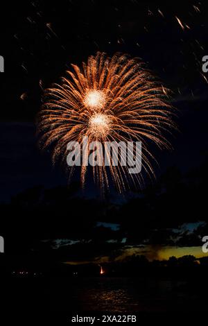 Colorful fireworks burst over water at night, with a dazzling orange and black firework in the sky Stock Photo