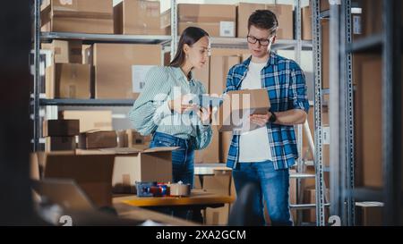 Female Sales Manager Using Tablet Computer, Talking to a Worker Holding a Cardboard Package, They Discuss Work and Smile. Warehouse with Parcels Ready for Shipment in the Background. Stock Photo