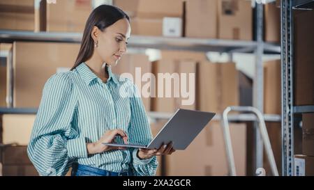 Portrait of a Worker Checking Inventory, Writing Down Notes on Laptop Computer. Young Woman Working in a Warehouse Storeroom with Rows of Shelves with Parcels, Packages with Orders Ready for Shipment. Stock Photo