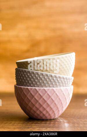 A close-up photo of three ceramic bowls stacked on a wooden tabletop. The bowls are different colors and textures, with the top bowl being a cream col Stock Photo