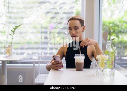 Portrait of beautiful Asian queer LGBT community supporter man with mustache wearing lipstick and jumpsuit using mobile phone in coffee shop Stock Photo