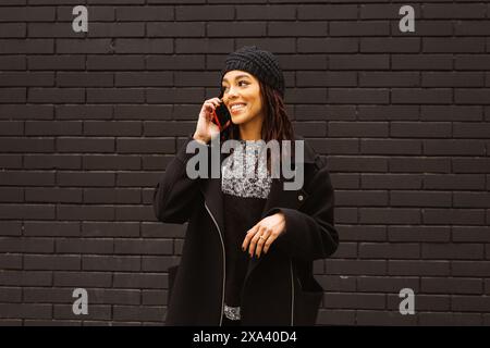 Young woman with dreadlocks smiling while talking on a red smartphone standing against a black brick wall on the street. Stock Photo