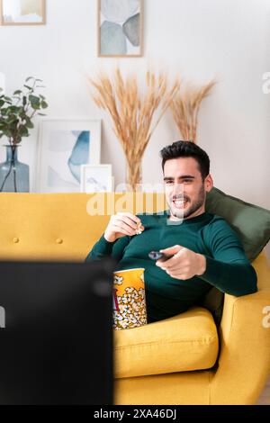 Man relaxing on a yellow sofa watching TV with remote and popcorn. Stock Photo