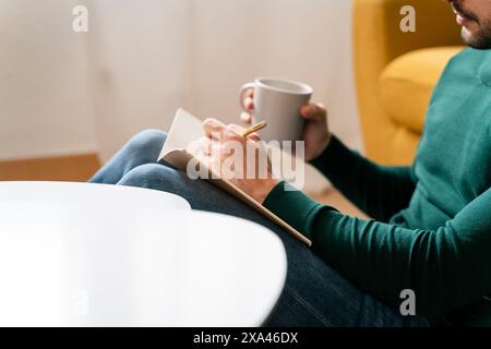 Person writing in notebook while holding a mug. Stock Photo