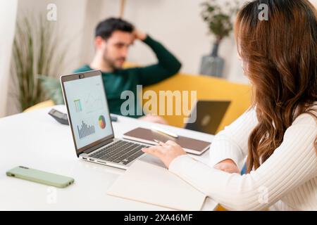 Woman working on laptop with charts as man relaxes in background Stock Photo