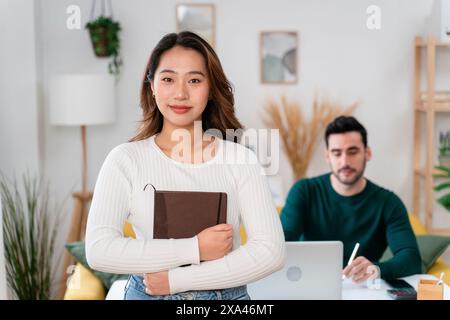 Young woman with a book standing in an office, man working on laptop behind her. Stock Photo