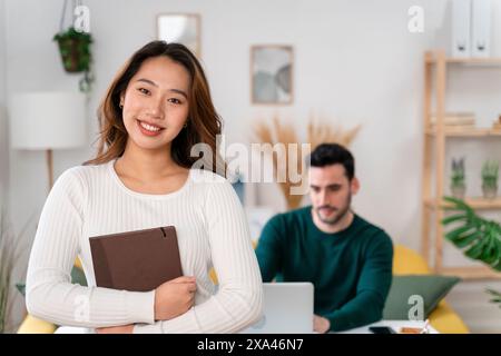 Smiling woman holding a notebook with a man working on a laptop in the background. Stock Photo