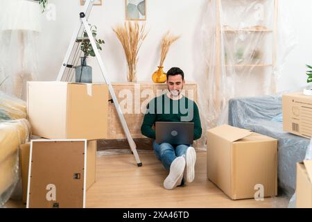 Man with laptop sitting on floor among moving boxes. Stock Photo