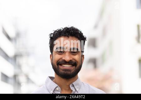 A cheerful man with a beard and curly hair is smiling at the camera in front of a blurred urban background. Stock Photo