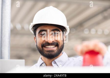 A smiling man in a white safety helmet is pressing a red button, with a steel structure in the background. Stock Photo
