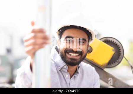 A smiling man in a white hard hat leans on a metal pole at a construction site. Stock Photo