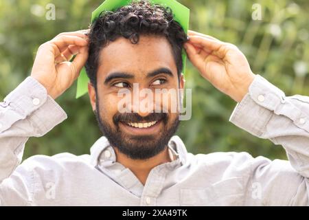 A smiling man with curly hair is putting on a green paper hat in a natural outdoor setting. Stock Photo