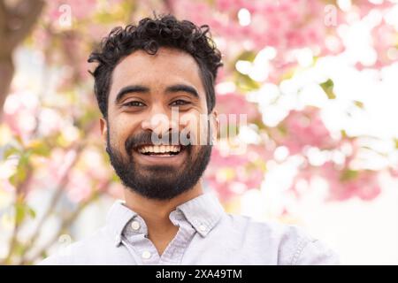 A smiling man with a beard and curly hair poses in front of a blooming pink flower tree. Stock Photo